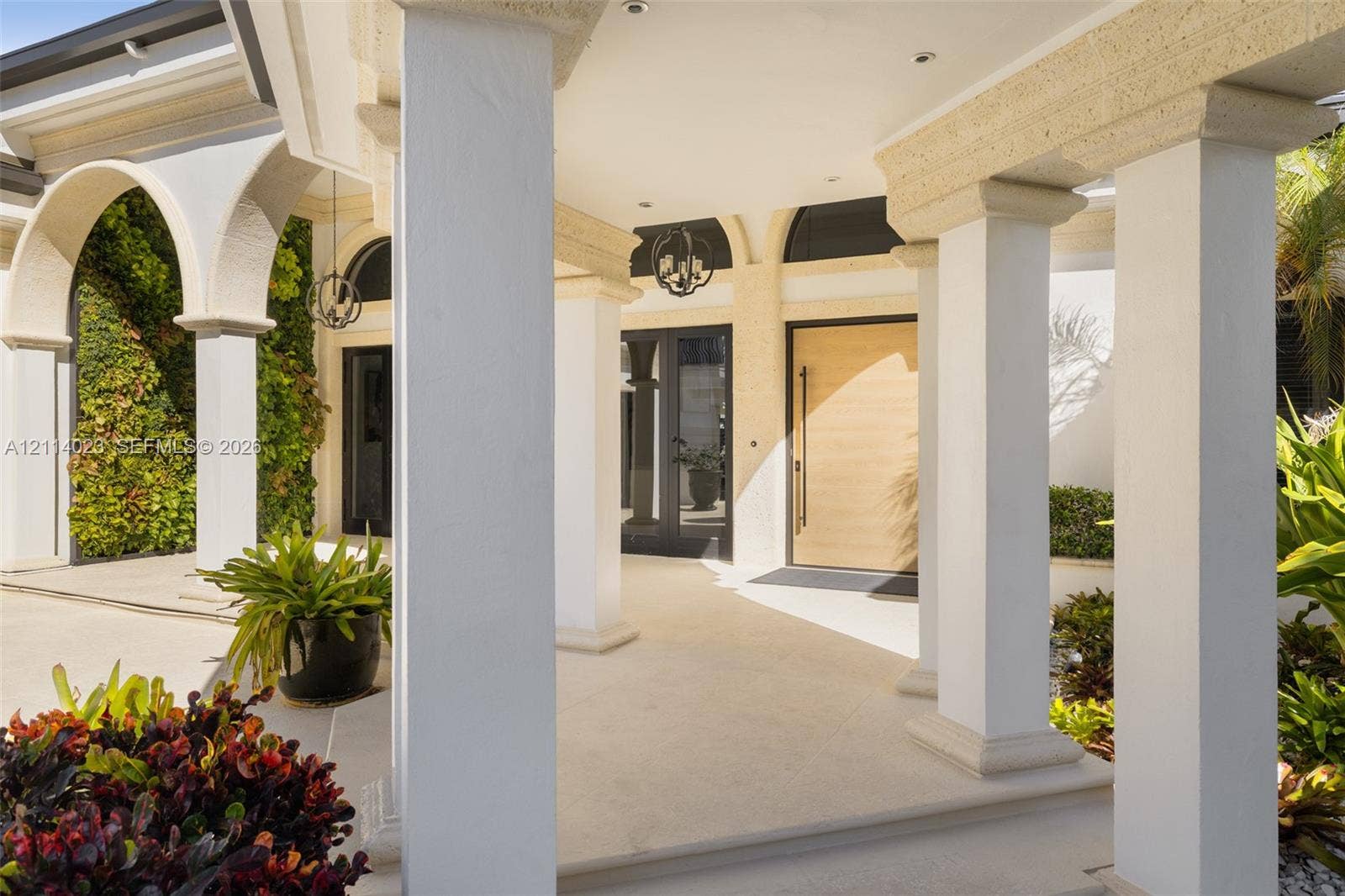 Covered portico featuring structural columns, arched architectural detailing, a natural wood-finish front door, recessed lighting, and integrated wall-climbing greenery