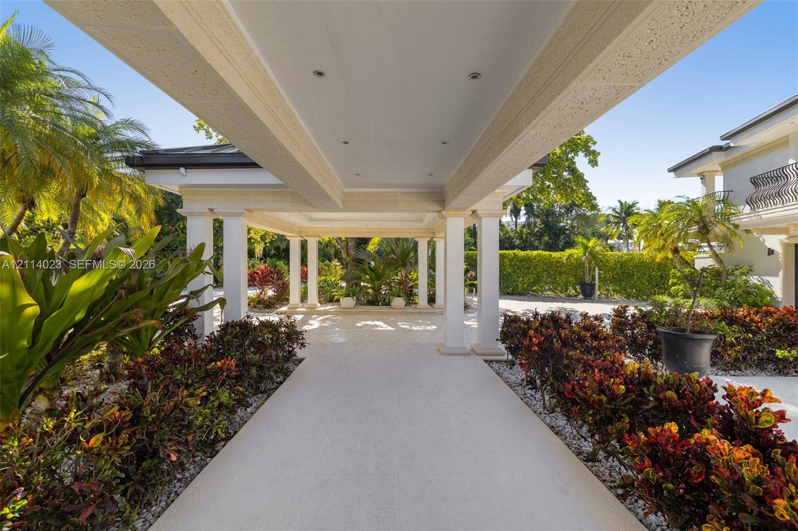 Covered porte-cochre featuring a light-toned stone tile surface, structural white columns, recessed lighting, and surrounding professional landscaping