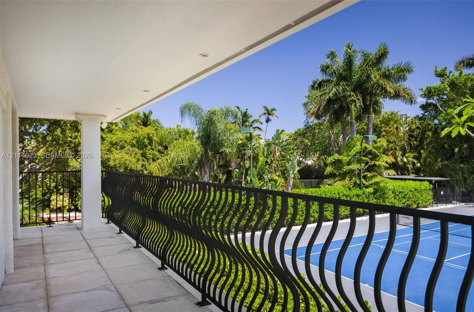 Covered balcony featuring stone-tile flooring, recessed lighting, and black metal wavy-pattern railing overlooking a hard-surface sport court and mature landscaping