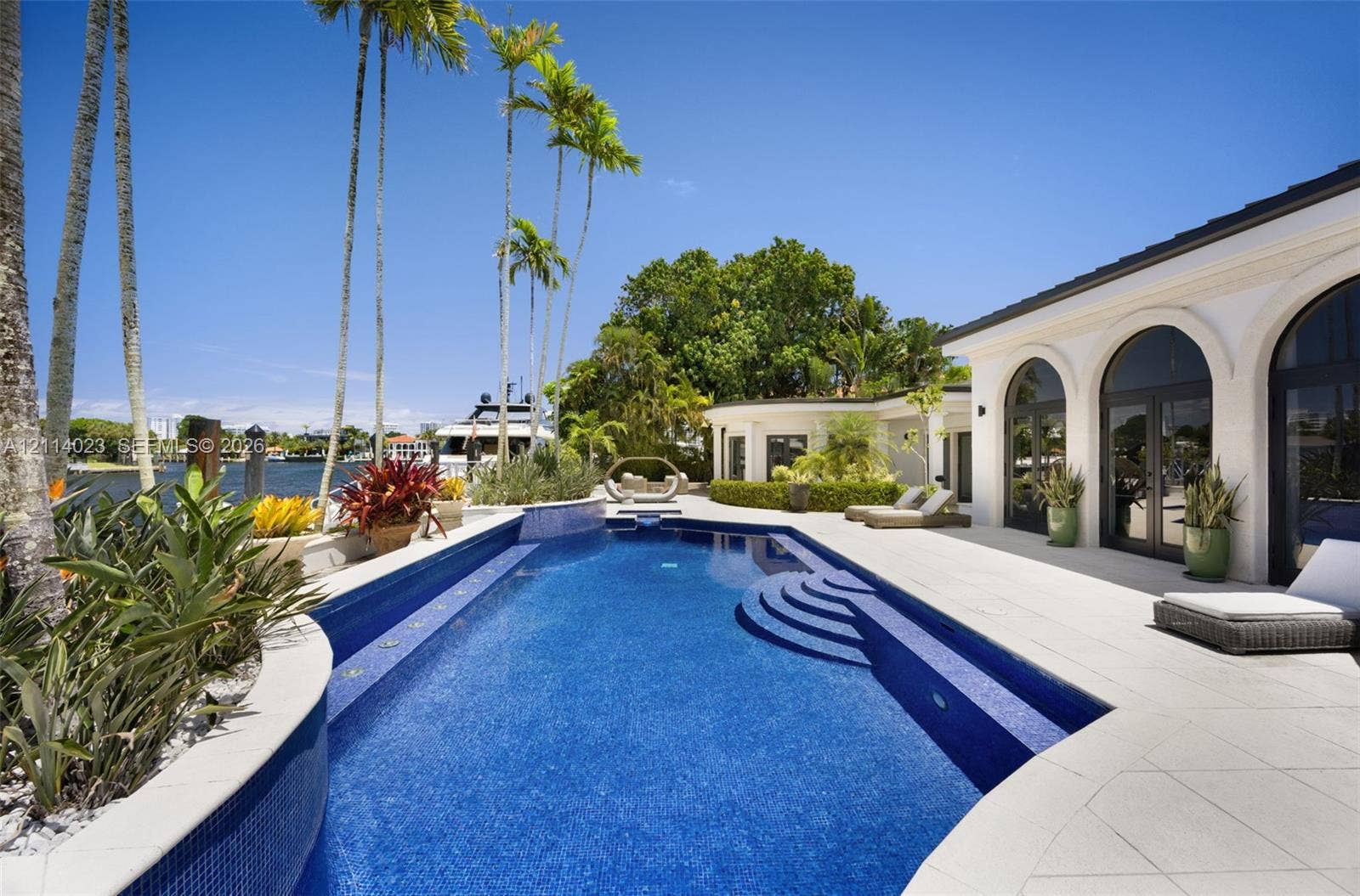 Waterfront pool area featuring a custom tiled swimming pool, built-in sun shelf, light-toned stone decking, arched glass entryways, and manicured tropical landscaping