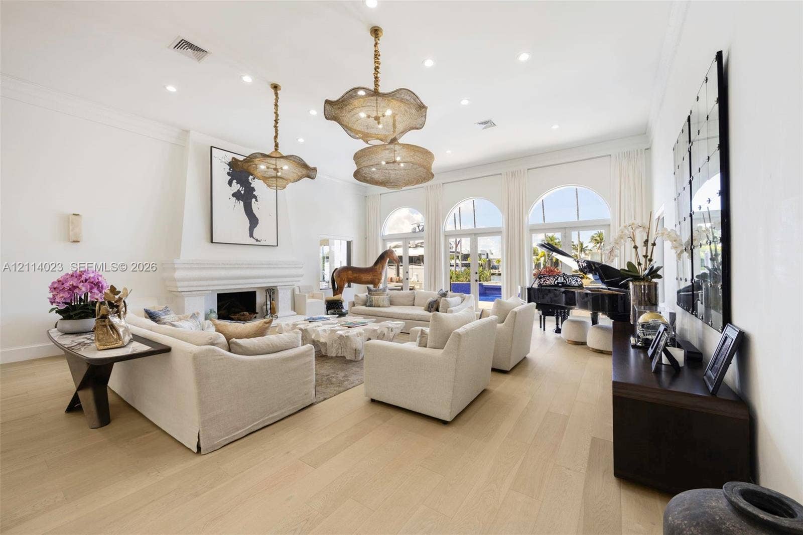 Expansive living area featuring high ceilings, wood-finish flooring, a prominent fireplace with a decorative mantel, sculptural pendant lighting, and triple arched glass doors