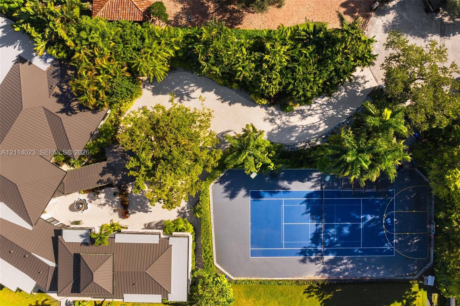 Aerial perspective of the estate featuring a multi-sport court with blue hard surfacing, expansive stone-paved driveway, metal roofing, and mature landscape canopy