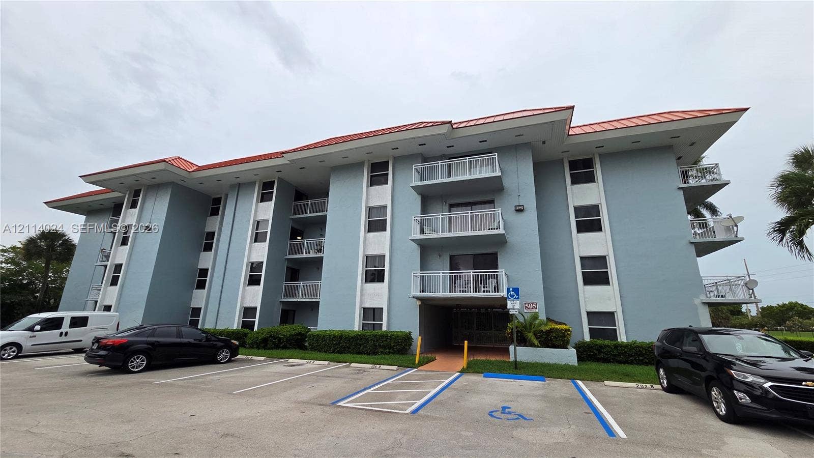 505 S Pine Island Road, #B205, Plantation, FL 33324 Multi-story residential building featuring a red metal roof, stucco exterior, stacked private balconies with railings, and paved surface parking