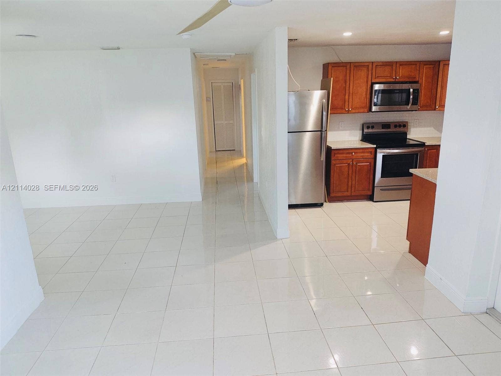 Kitchen featuring wood-finish cabinetry, neutral solid-surface countertops, and stainless steel appliances, set against white tile flooring