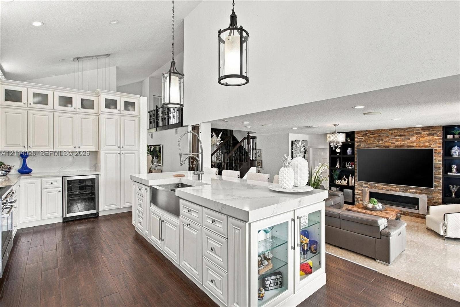 5257 SW 158th Avenue, Miramar, FL 33027 Kitchen featuring white shaker-style cabinetry, an expansive center island with an integrated farmhouse sink, glass-front display shelving, stone-veneer fireplace surround, and dark wood-finish flooring