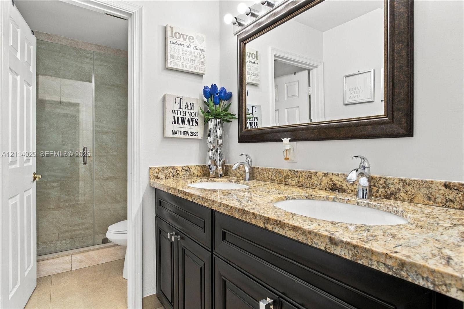 5257 SW 158th Avenue, Miramar, FL 33027 Dual-sink vanity with stone countertop, dark-toned cabinetry, framed wall mirror, multi-bulb vanity light, and tiled walk-in shower with glass enclosure