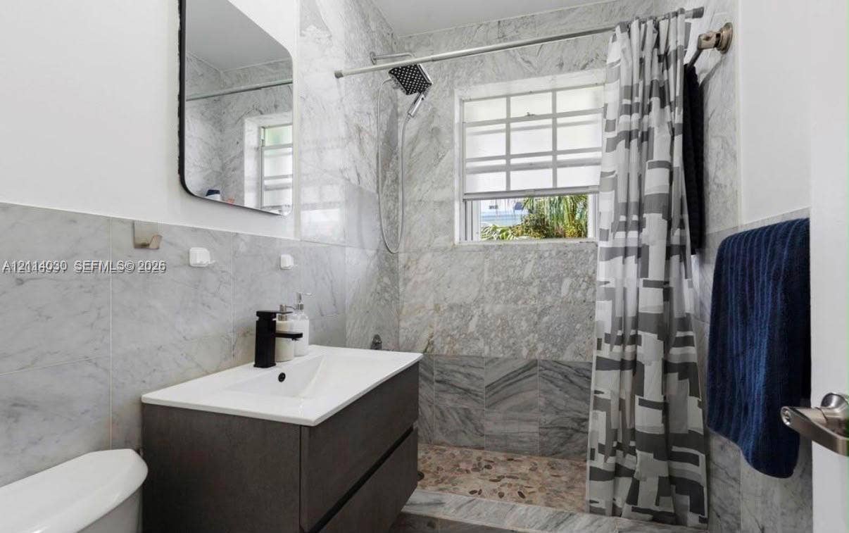 8550 Byron Avenue, #2H, Miami Beach, FL 33141 Bathroom featuring floor-to-ceiling stone tile, floating vanity with integrated sink, matte black faucet, and rainfall shower head