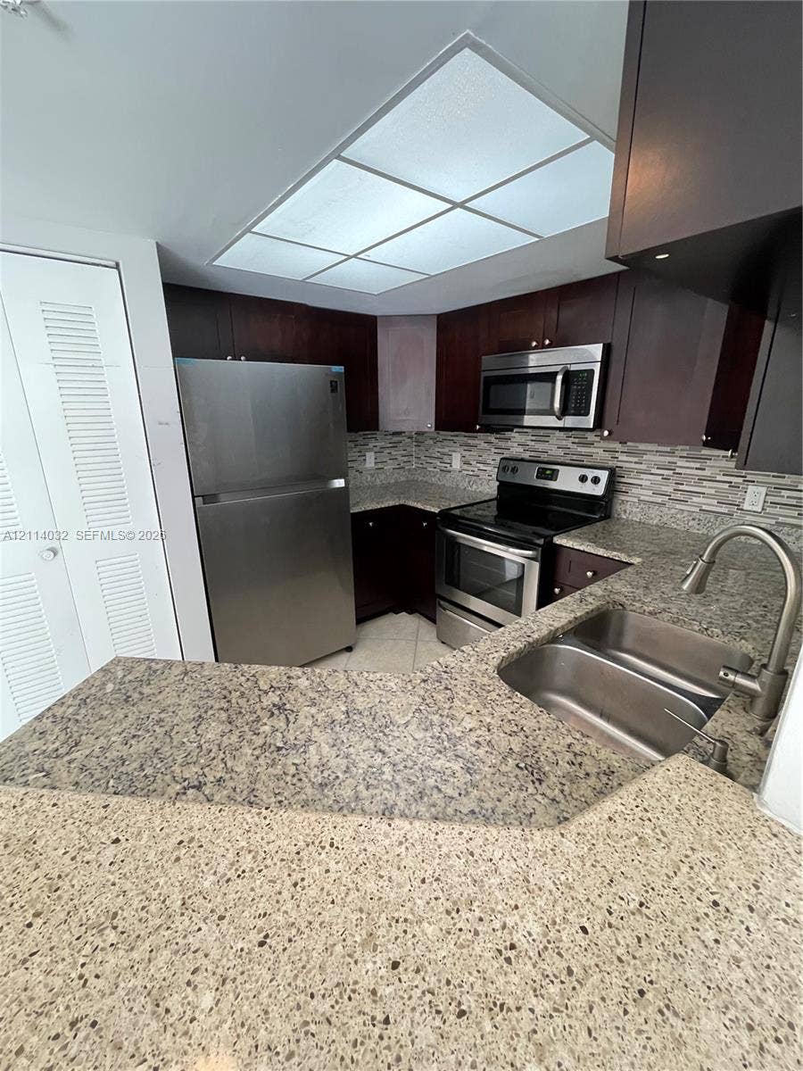 4177 N Haverhill Road, #821, West Palm Beach, West Palm Beach, FL 33417 Kitchen featuring dark wood-finish cabinetry, speckled stone-composite countertops, stainless steel appliances, a multi-textured linear tile backsplash, and a double-basin stainless steel sink with a pull-down faucet