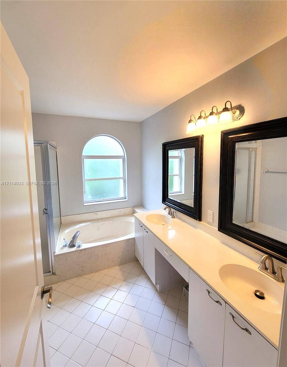 683 NW 88th Drive, Coral Springs, Coral Springs, FL 33071 Bathroom featuring a dual-sink vanity with a solid-surface countertop, an arched window above a soaking tub with tile surround, a separate glass-enclosed shower, and tile flooring