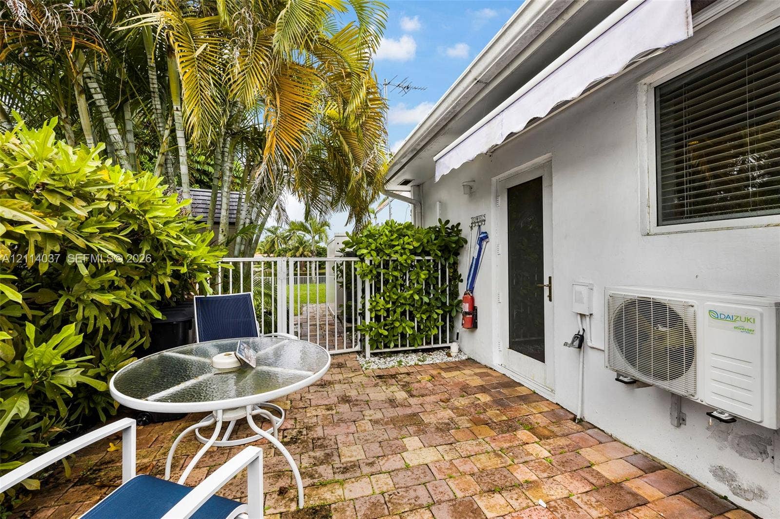 8436 SW 143rd Avenue, Miami, FL 33183 Private patio featuring brick pavers, a retractable awning, and mature palm landscaping with a metal perimeter fence