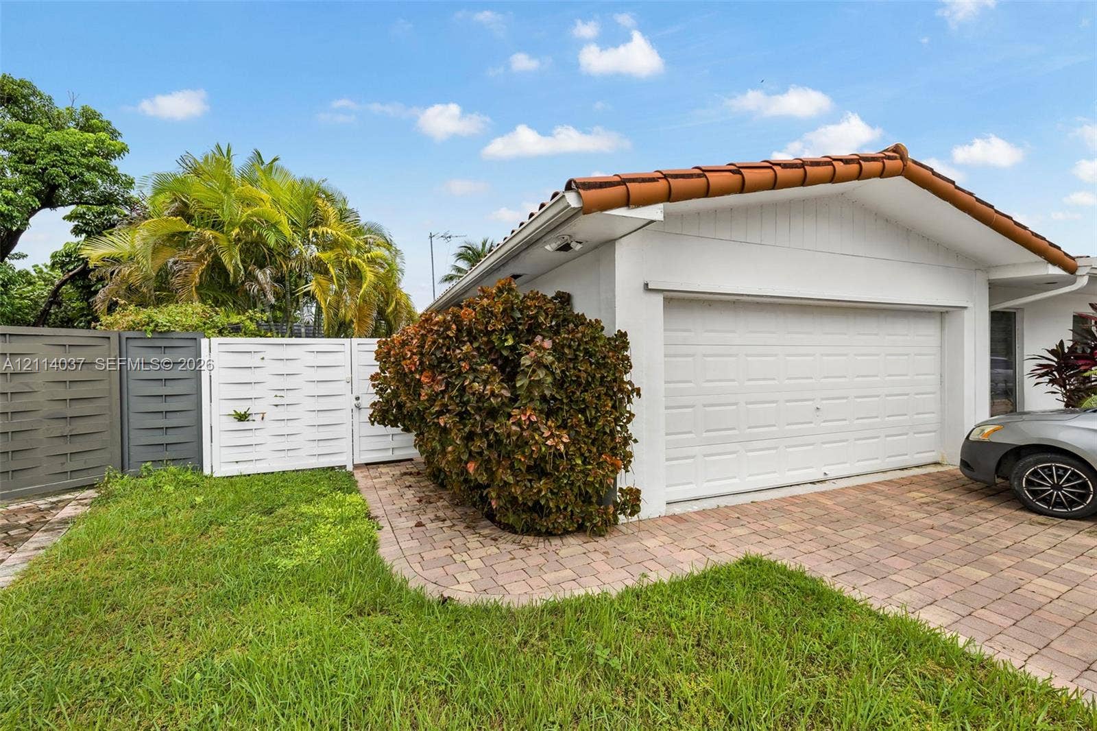 8436 SW 143rd Avenue, Miami, FL 33183 Exterior of a structure featuring a tiled roof, multi-panel garage door, and brick-paver driveway, with white privacy fencing and perimeter landscaping