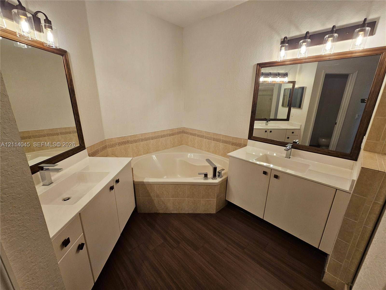 Dual vanities with rectangular sinks and white cabinetry, wood-finish flooring, corner soaking tub, and framed wall mirrors with linear lighting