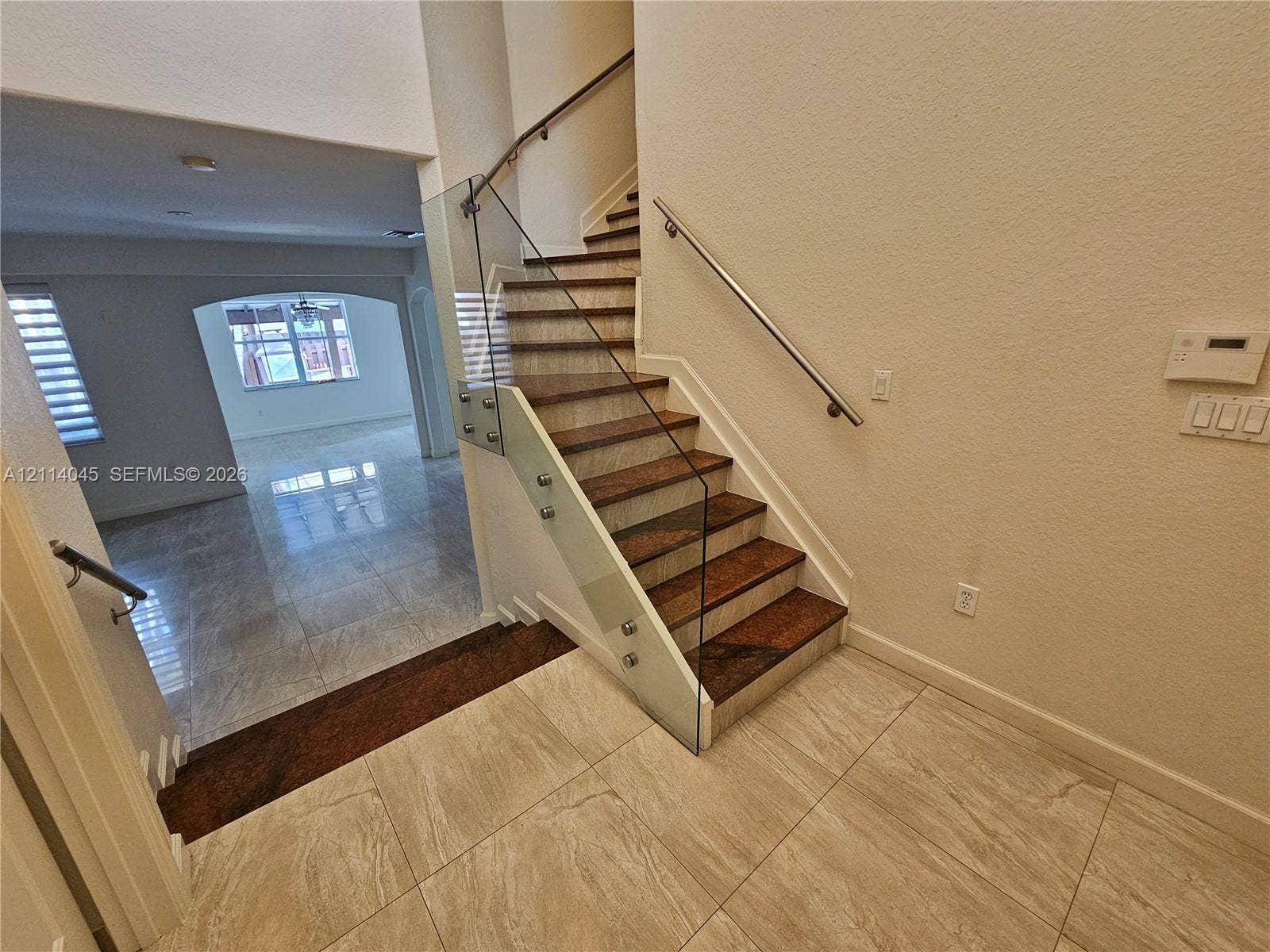 Staircase featuring wood-finish treads, glass panel railing with metallic standoffs, and a wall-mounted metal handrail, complemented by neutral tile flooring