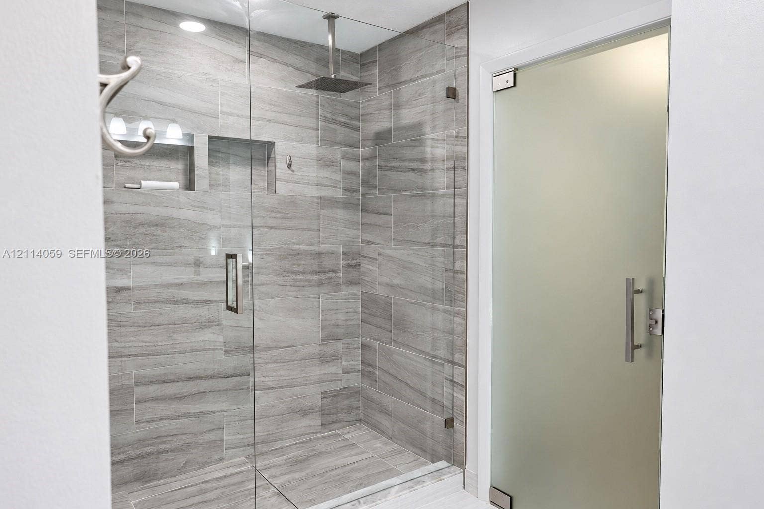 8023 N Tranquility Lake Drive, Delray Beach, FL 33446 Walk-in shower featuring floor-to-ceiling stone-pattern tile, a ceiling-mounted rainfall showerhead, recessed shelving, and a frameless glass enclosure with a frosted glass entry door