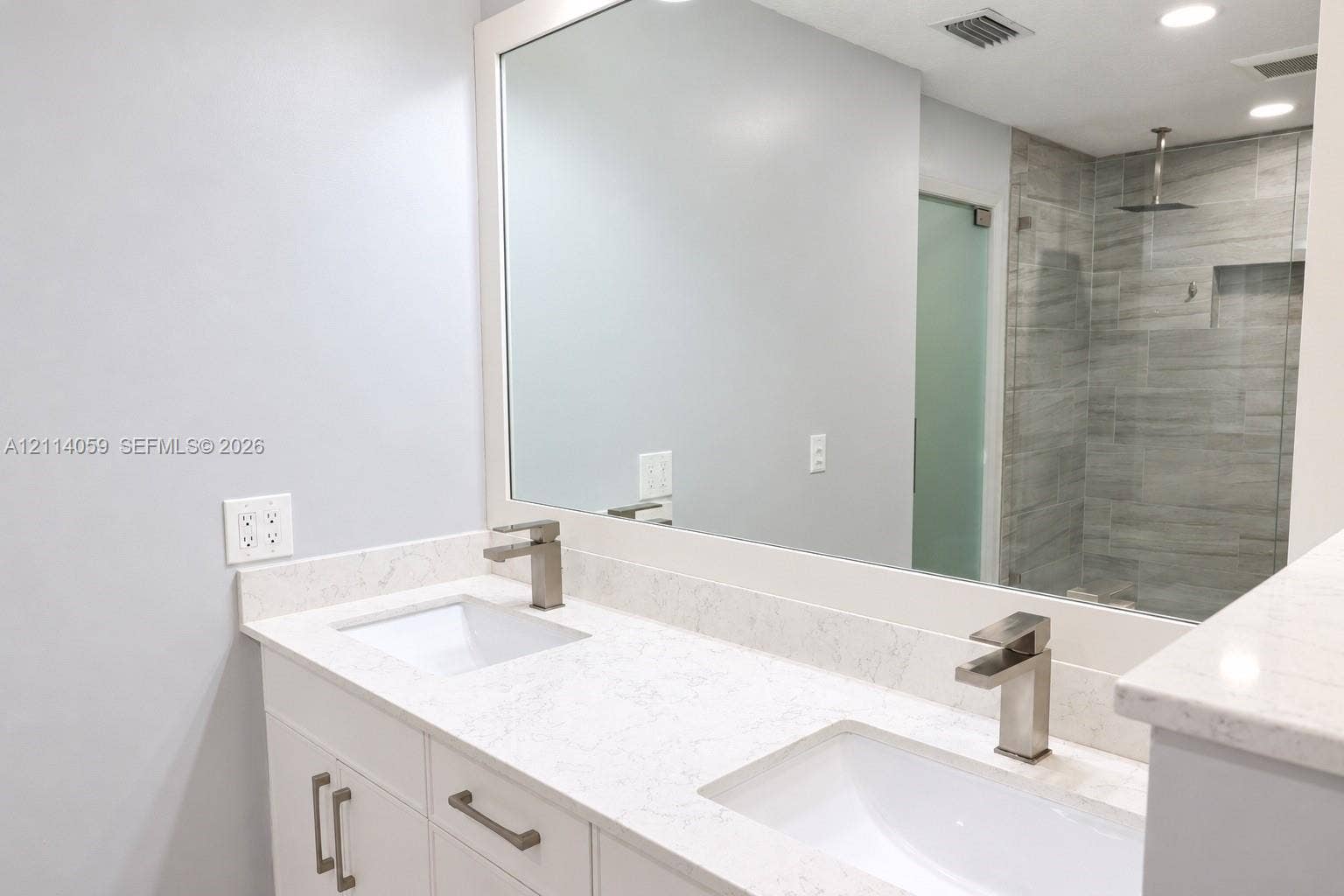 8023 N Tranquility Lake Drive, Delray Beach, FL 33446 Double vanity featuring white cabinetry, stone-finish countertops, rectangular undermount sinks, and brushed-finish faucets, paired with a walk-in shower encased in glass with tile wall surfacing and a built-in recessed shelf