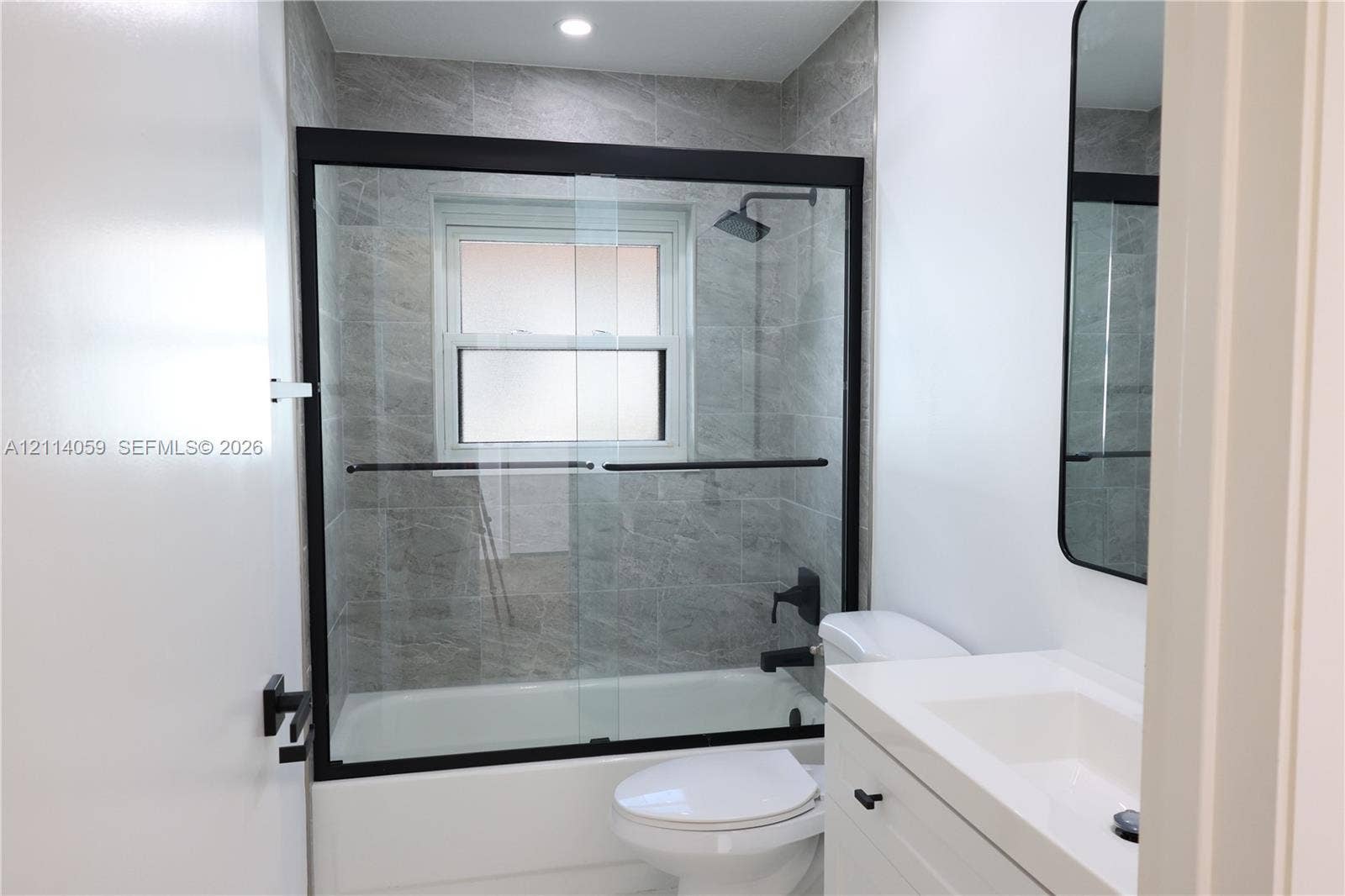 8023 N Tranquility Lake Drive, Delray Beach, FL 33446 Bathtub enclosure with glass sliding doors, matte black fixtures, full-height stone-finish tile surround, recessed lighting, and a white vanity with an integrated sink