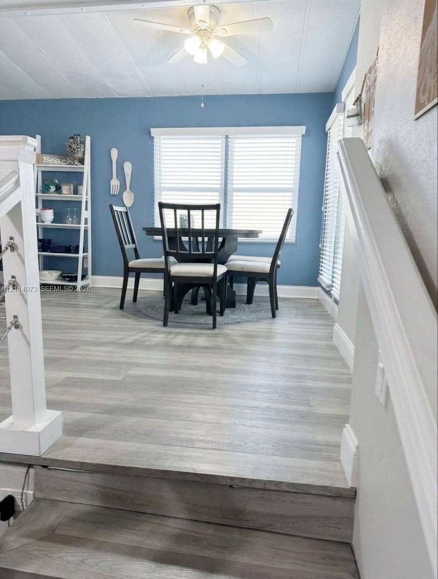 Wood-finish flooring, white ceiling fan with integrated lighting, dual windows with horizontal blinds, and a white stair railing with cable accents
