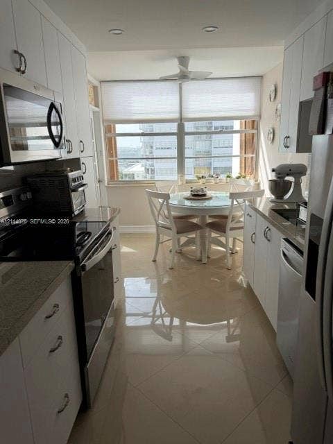 Galley kitchen featuring white shaker-style cabinetry, stone countertops, stainless steel appliances, large-format tile flooring, and oversized windows with window treatments