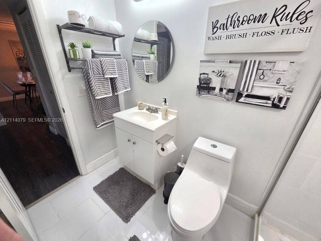 9241 Sunrise Lakes Boulevard, #109, Sunrise, FL 33322 White vanity with integrated sink, circular wall-mounted mirror, white tile flooring, and two-piece elongated toilet