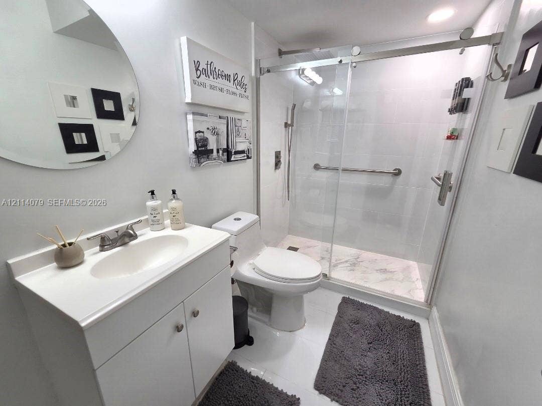 9241 Sunrise Lakes Boulevard, #109, Sunrise, FL 33322 White vanity with integrated basin, walk-in shower featuring a glass sliding door, stone-patterned shower floor tile, and a circular wall-mounted mirror
