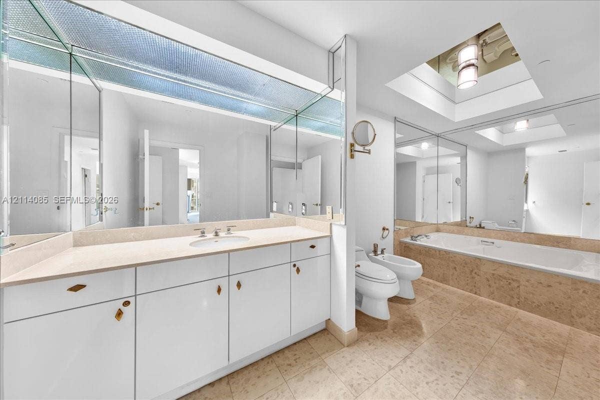 10101 Collins Avenue, #8D, Bal Harbour, FL 33154 Bathroom featuring a white vanity with integrated stone countertop and sink, large wall-to-wall mirror, skylight, soaking tub with stone surround, and tile flooring