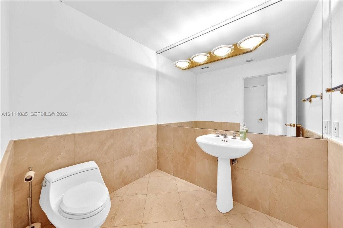 10101 Collins Avenue, #8D, Bal Harbour, FL 33154 Pedestal sink with polished metal faucet, full-width wall mirror, four-bulb vanity light fixture, and large-format tile flooring and wainscoting