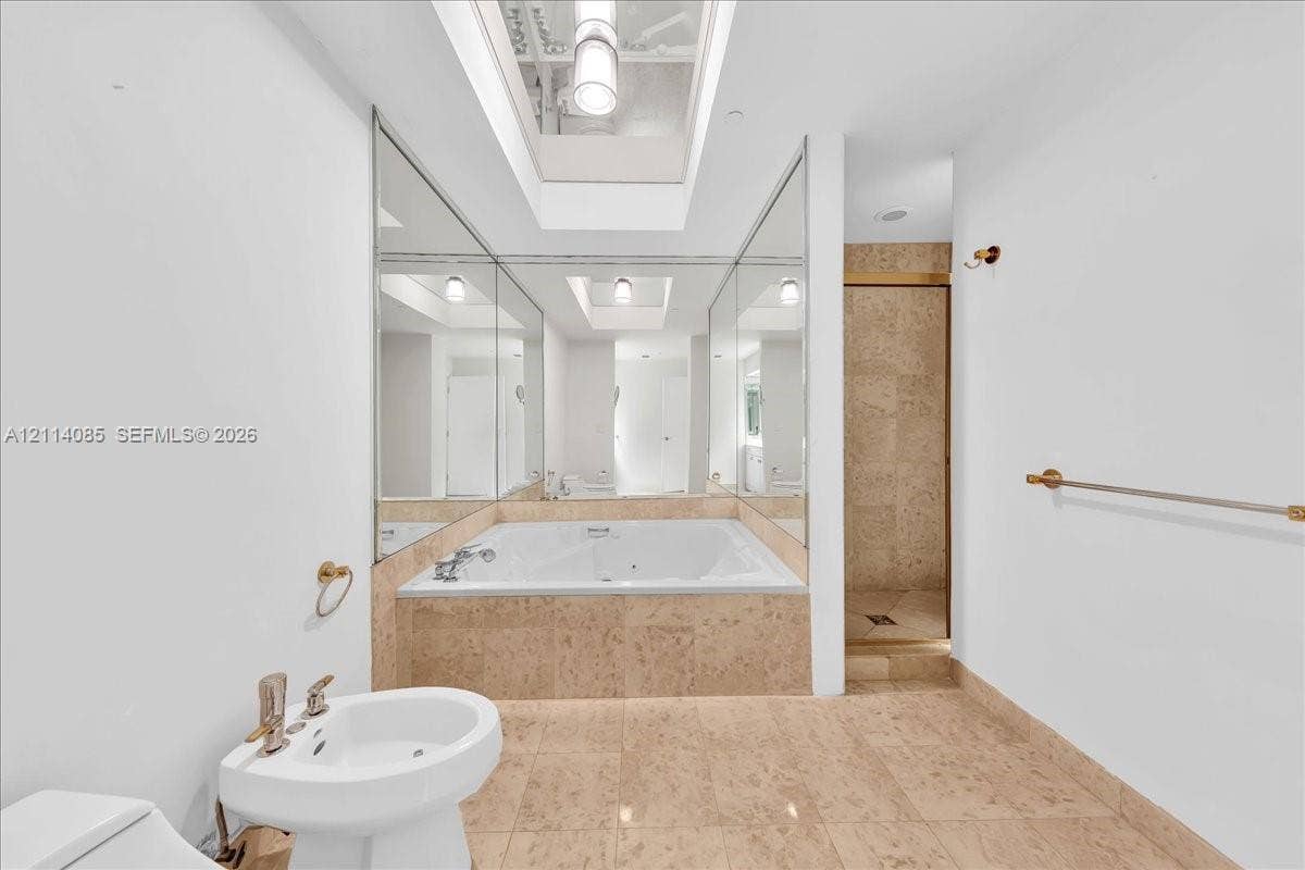 10101 Collins Avenue, #8D, Bal Harbour, FL 33154 Jetted soaking tub with stone tile surround, skylight, wall-to-wall mirrors, pedestal sink, and separate walk-in shower with stone tile wall finishes