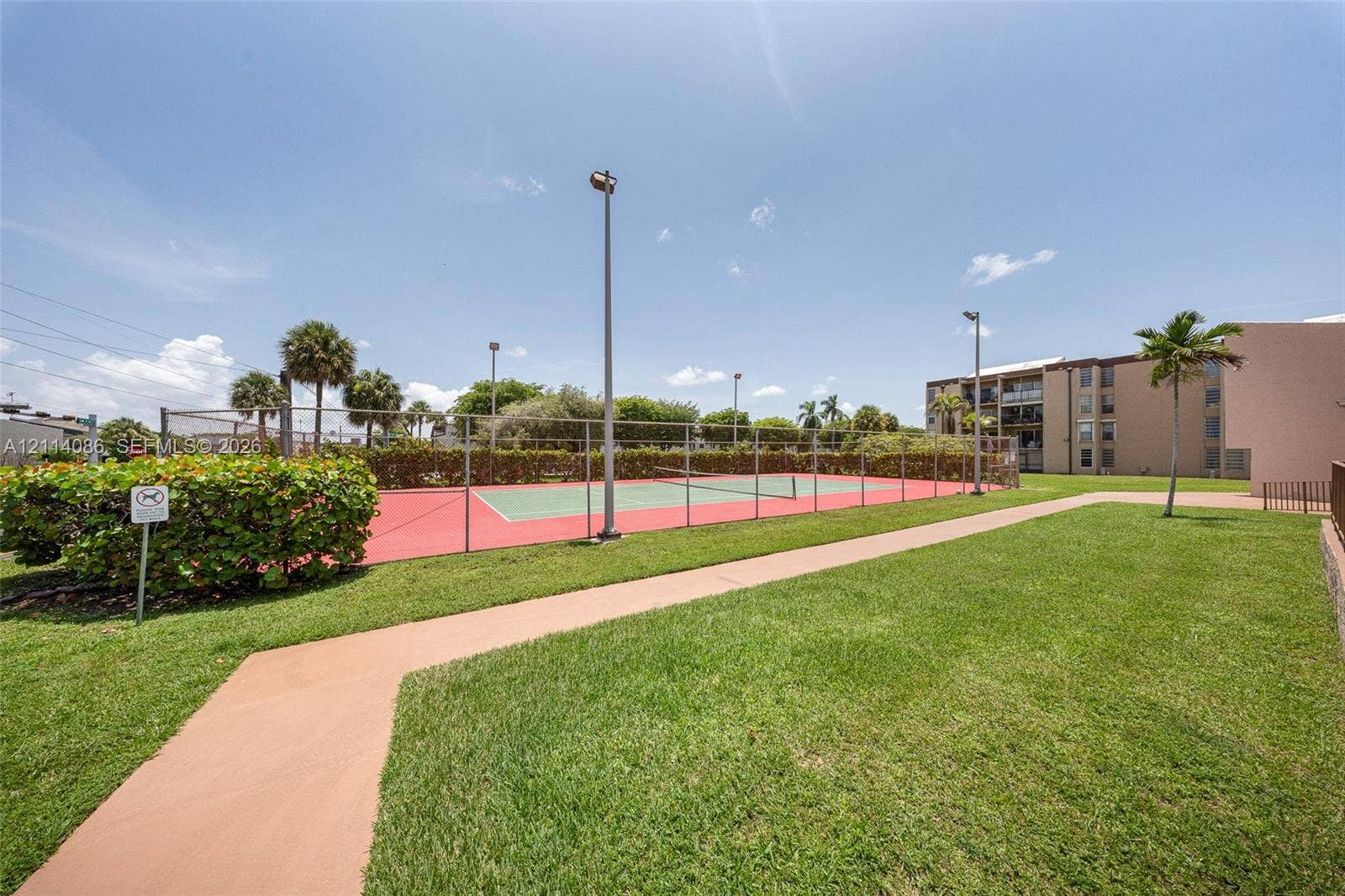 706 NW 87th Avenue, #204, Miami, Miami, FL 33172 Fenced tennis court with a red and green surface, surrounding perimeter lighting, and paved walkway set within a landscaped grass lawn