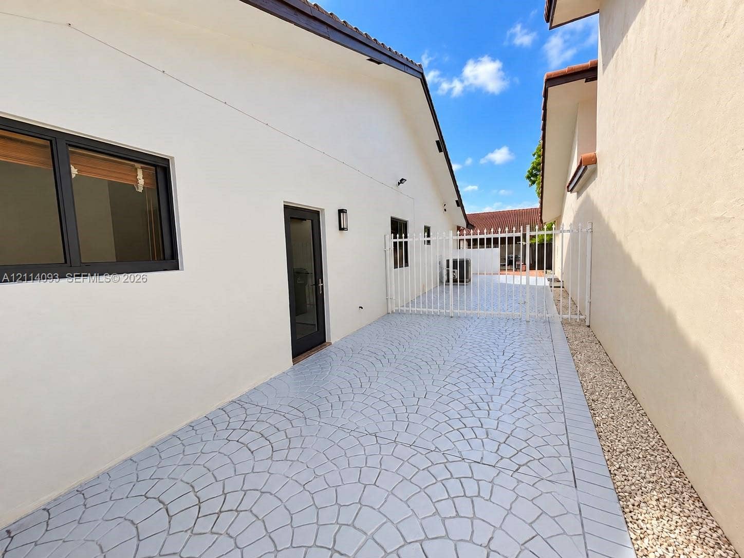 4916 SW 142nd Place, #4916, Miami, Miami, FL 33175 Side exterior featuring decorative stamped concrete paving, white metal perimeter fencing, wall-mounted light fixtures, and a glass-paneled entry door