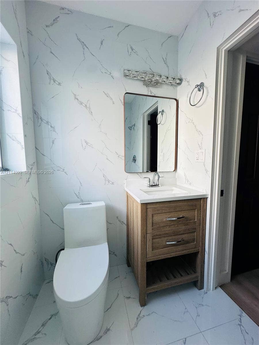 3052 SW 6th Street, Miami, FL 33135 Full-height stone-patterned wall tile, wood-finish vanity with two drawers and lower open shelving, integrated white countertop with rectangular sink, polished chrome faucet, and rectangular mirror