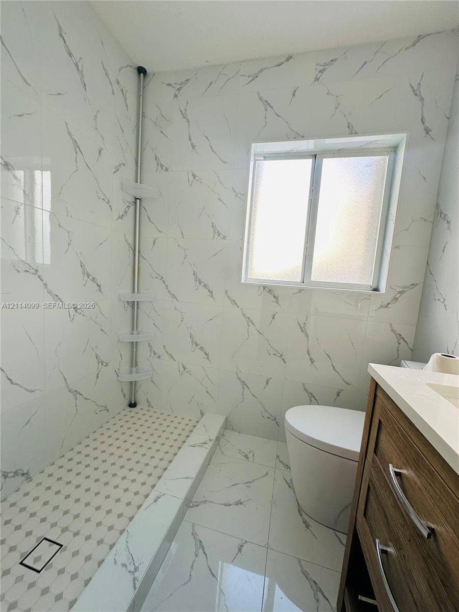 3052 SW 6th Street, Miami, FL 33135 Walk-in shower featuring floor-to-ceiling large-format tile with grey veining, a geometric mosaic tile shower floor, a light-toned wood-finish vanity with a solid-surface countertop, and a sliding window