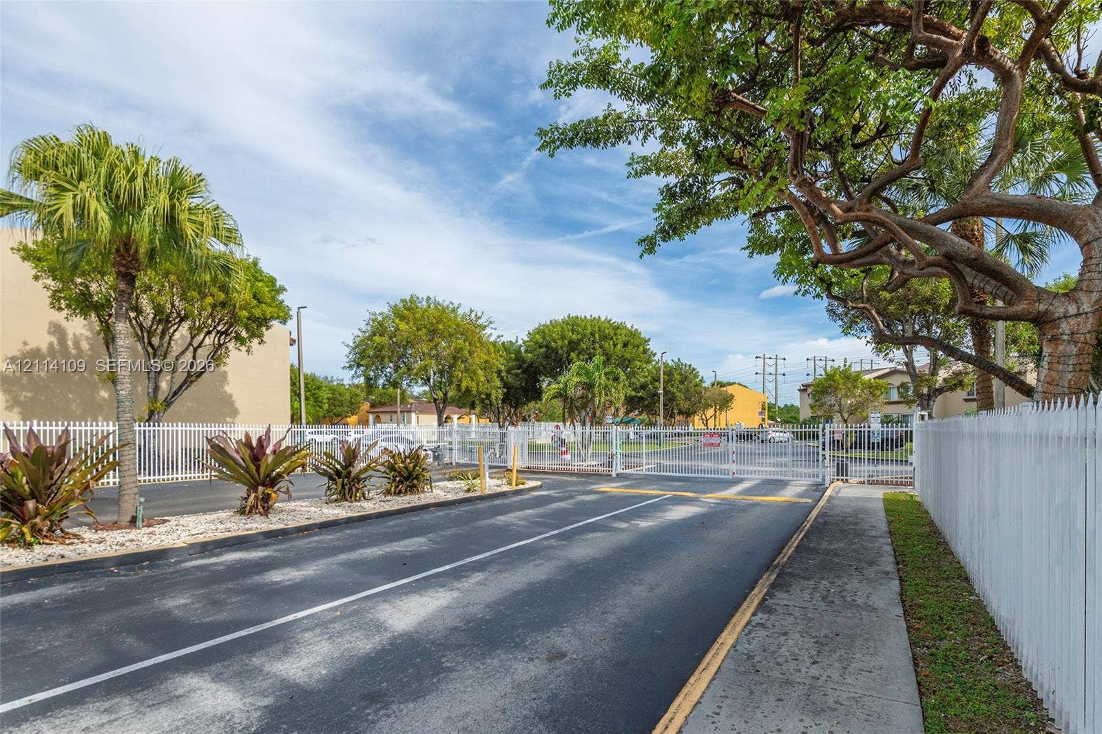 15761 SW 137th Avenue, #104, Miami, Miami, FL 33177 Gated entryway featuring a paved access road, white perimeter fencing, and mature landscaping