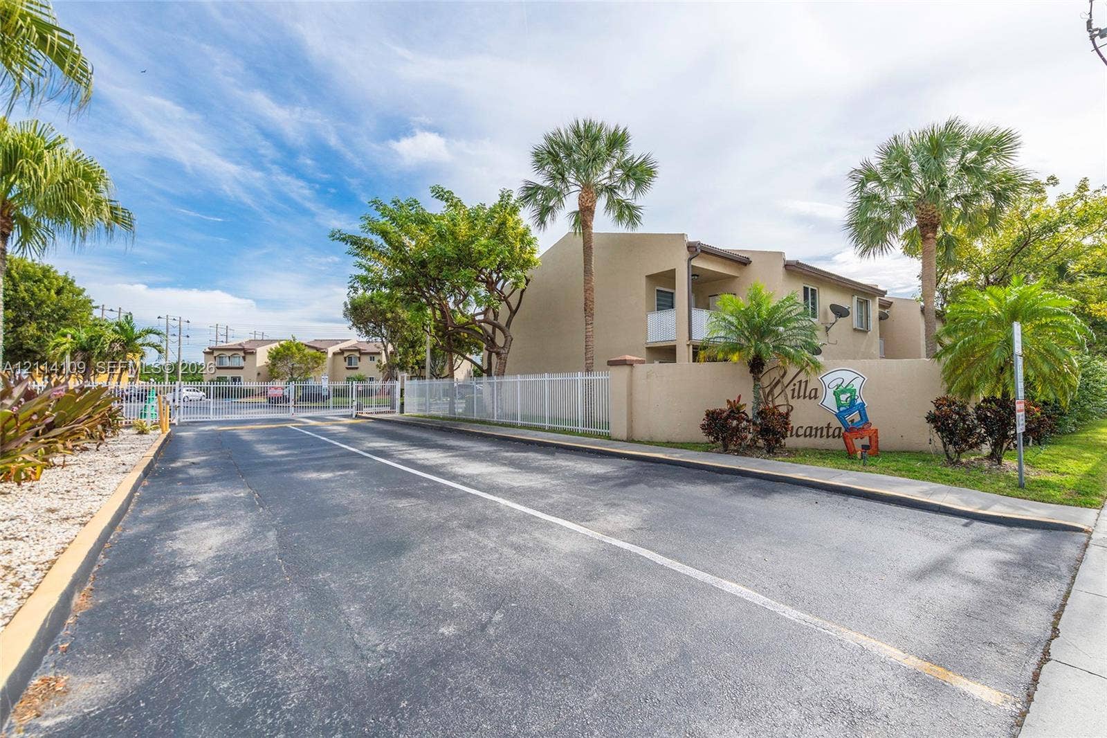 15761 SW 137th Avenue, #104, Miami, Miami, FL 33177 Gated entrance featuring stucco exterior walls, paved driveway, white perimeter fencing, and mature palm landscaping