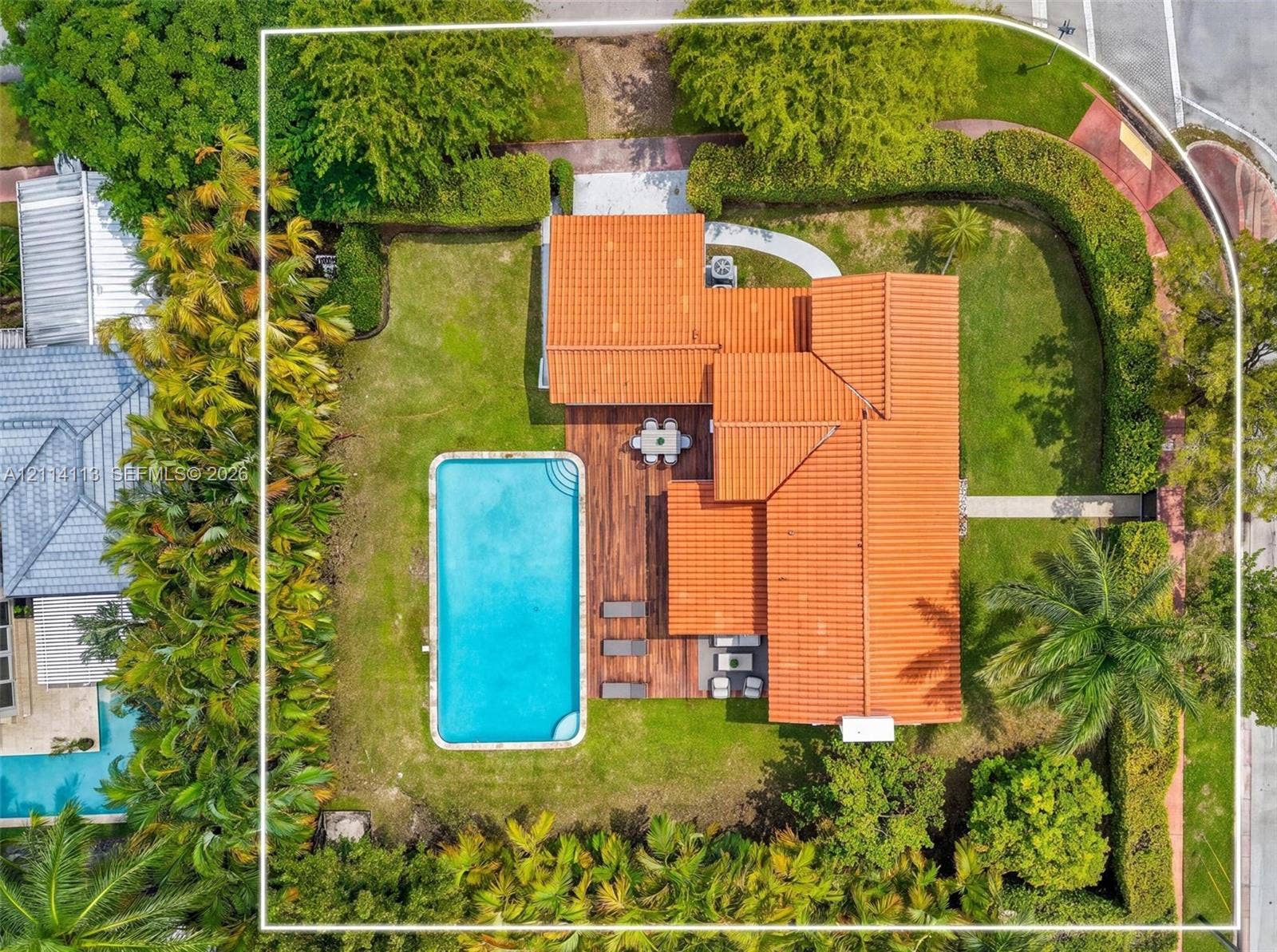 5386 Alton Road, Miami Beach, FL 33140 Aerial perspective of a corner lot property featuring a barrel tile roof, an in-ground swimming pool, wood-finish decking, and perimeter privacy hedging