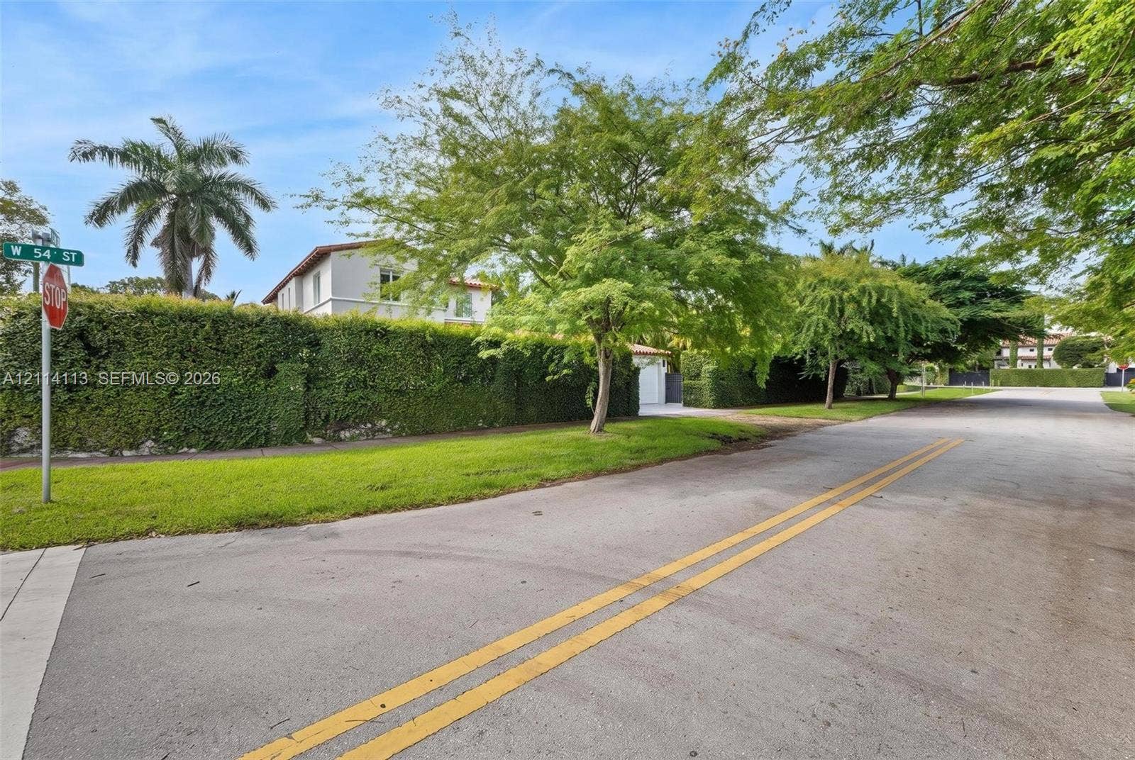 5386 Alton Road, Miami Beach, FL 33140 View of the side W 54th street featuring a tall perimeter privacy hedge, manicured lawn, and mature trees