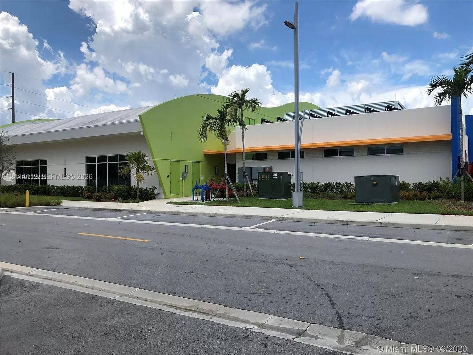 Modern commercial exterior featuring a contemporary curved lime green facade, large windows, and manicured perimeter landscaping with palm trees