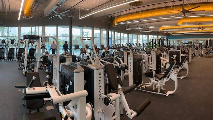 Expansive fitness center featuring floor-to-ceiling windows, high ceilings with exposed ductwork, rubberized flooring, and an extensive array of integrated exercise equipment