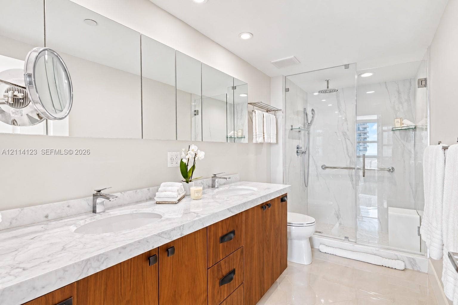 1155 Brickell Bay Drive, #2609, Miami, FL 33131 Double vanity featuring a light-toned stone countertop with dual undermount sinks, wood-finish cabinetry, floor-to-ceiling mirrored medicine cabinets, and a glass-enclosed shower with tiled walls and a built-in bench