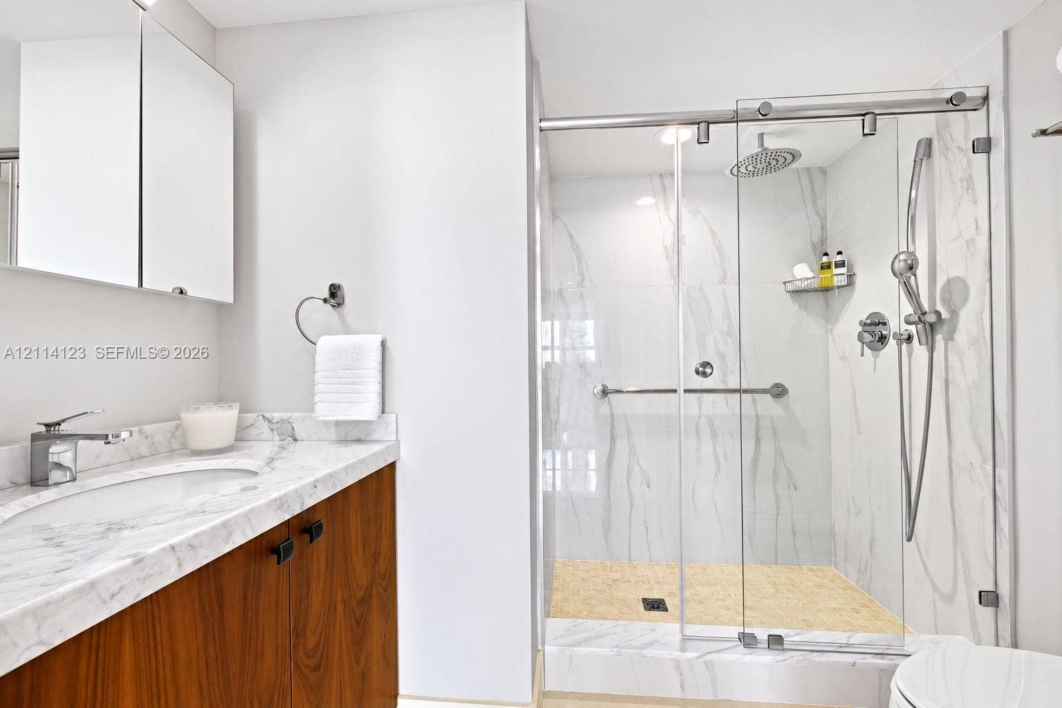 1155 Brickell Bay Drive, #2609, Miami, FL 33131 Bathroom featuring a wood-finish vanity with a stone-top integrated sink, stone-surround shower with a glass sliding door, rainfall showerhead, and a mirrored medicine cabinet