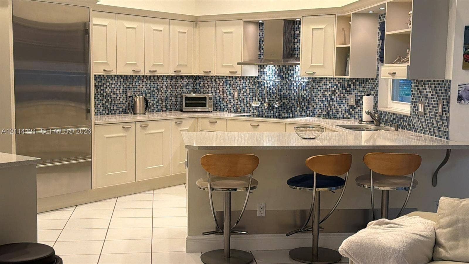 9012 Southern Orchard Road N, Davie, FL 33328 Kitchen featuring cream cabinetry, stone-finish countertops, full-height blue glass tile backsplash, stainless steel refrigerator, and stainless steel range hood