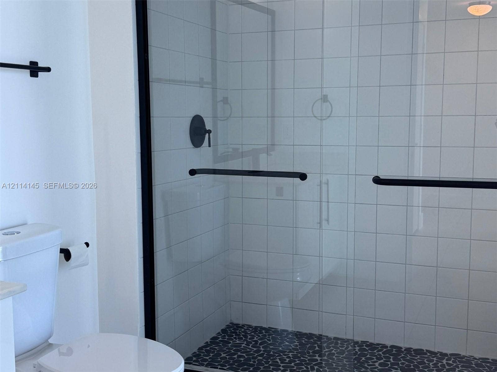 Walk-in shower featuring a pebble-stone floor, white square-tiled walls, black-framed glass enclosure, and matte black hardware