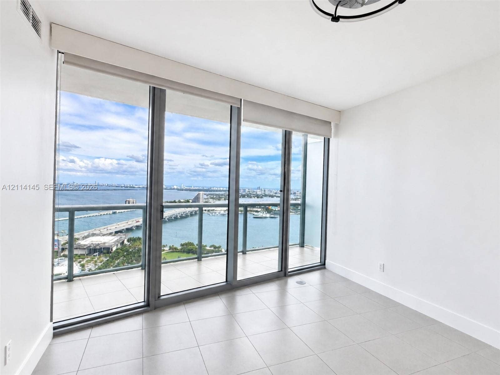 Floor-to-ceiling glass panels, expansive private balcony with metal railing, neutral tile flooring, and overhead circular light fixture