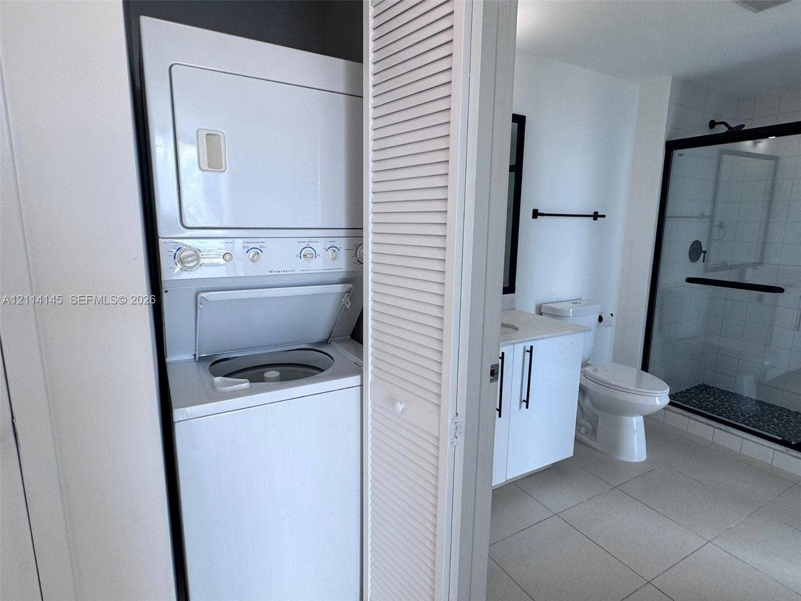 Stacked laundry unit with louvered closet doors, white vanity with stone-style countertop, vitreous china toilet, and walk-in shower with matte black hardware and tile flooring