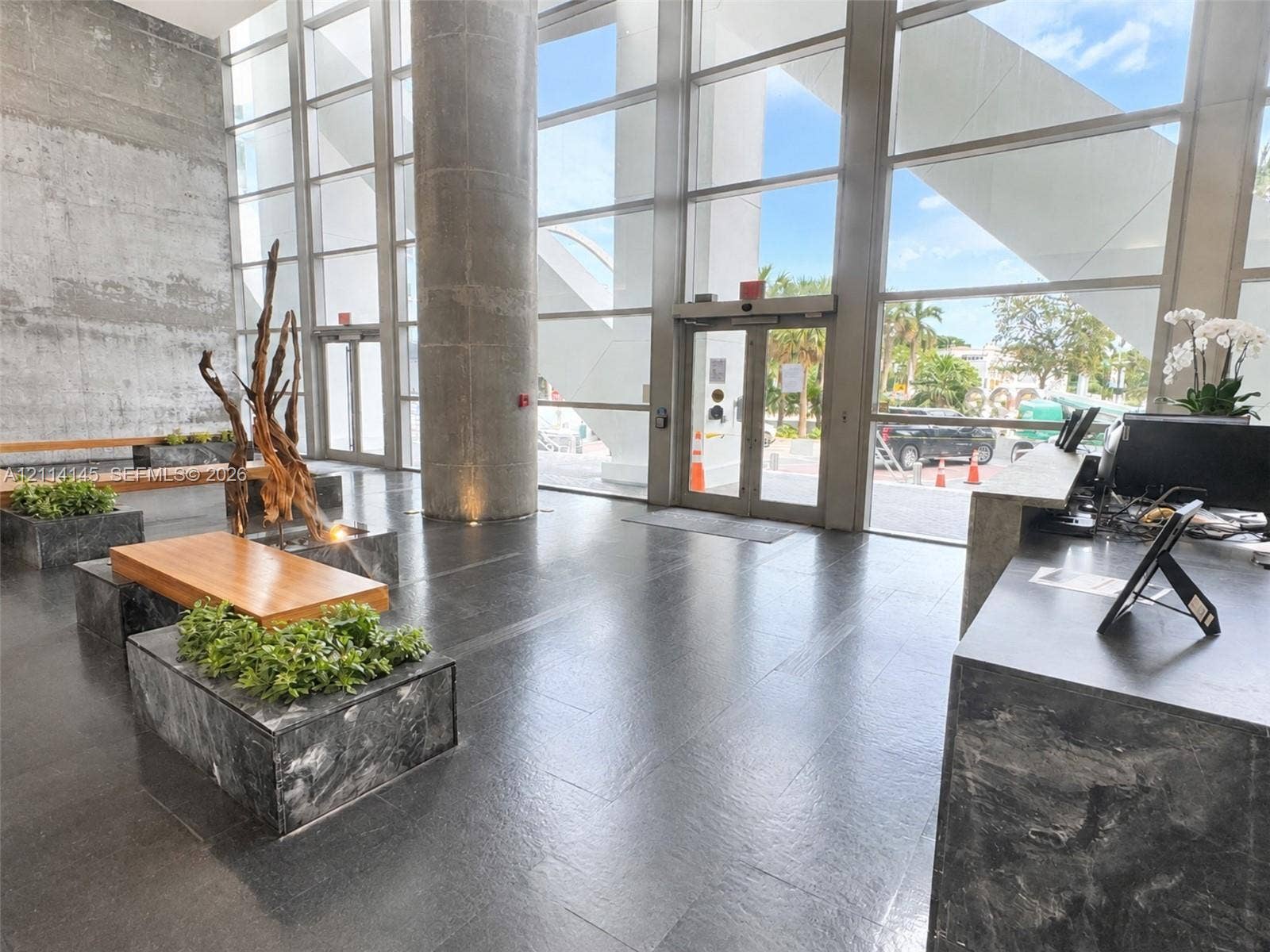 Double-height floor-to-ceiling windows, polished stone flooring, exposed concrete accent wall, integrated stone-clad planters, and architectural concrete column