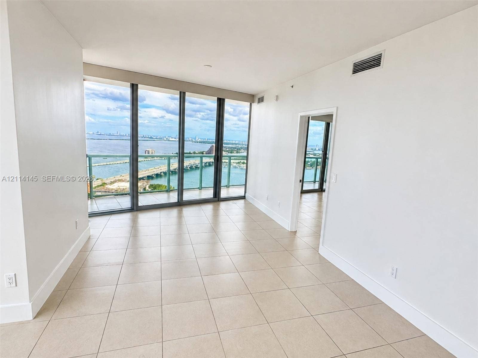 Floor-to-ceiling glass windows, ceramic tile flooring, and a private balcony featuring a glass railing with water and cityscape surroundings