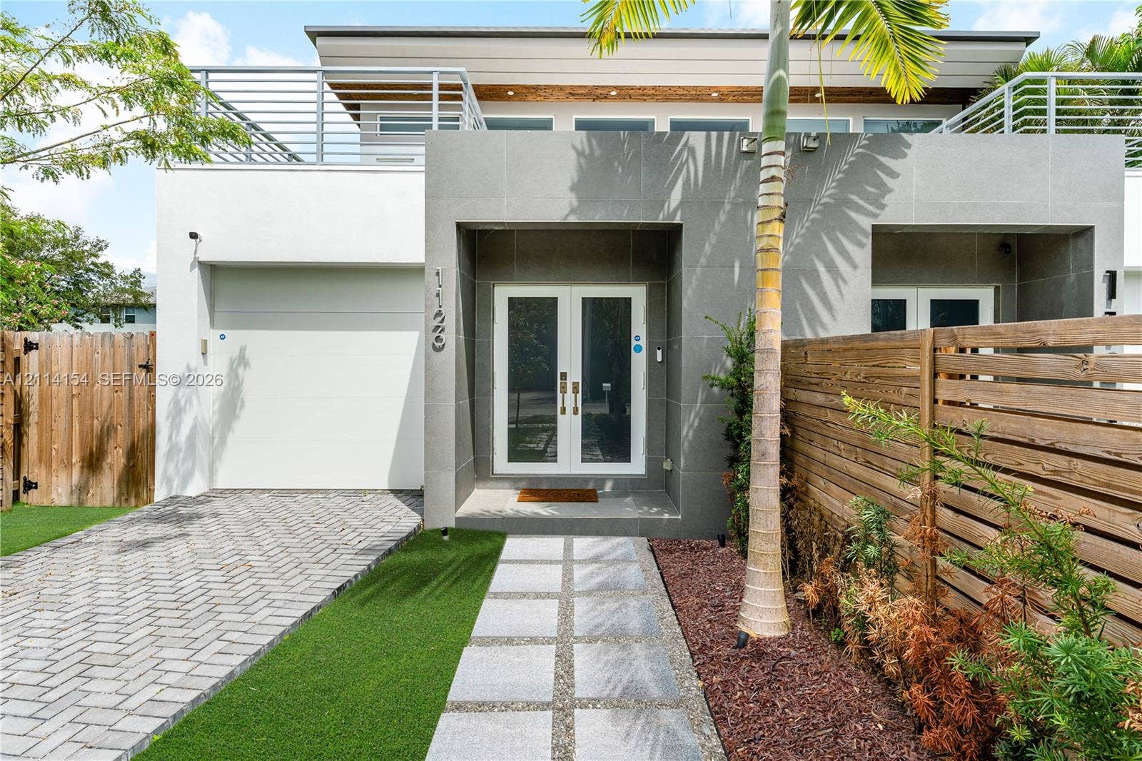 1126 NE 2nd Avenue, Fort Lauderdale, FL 33304 Modern exterior featuring a paved stone driveway, synthetic turf lawn, second-story balcony with metal railings, and a symmetrical glass-panel double door entrance