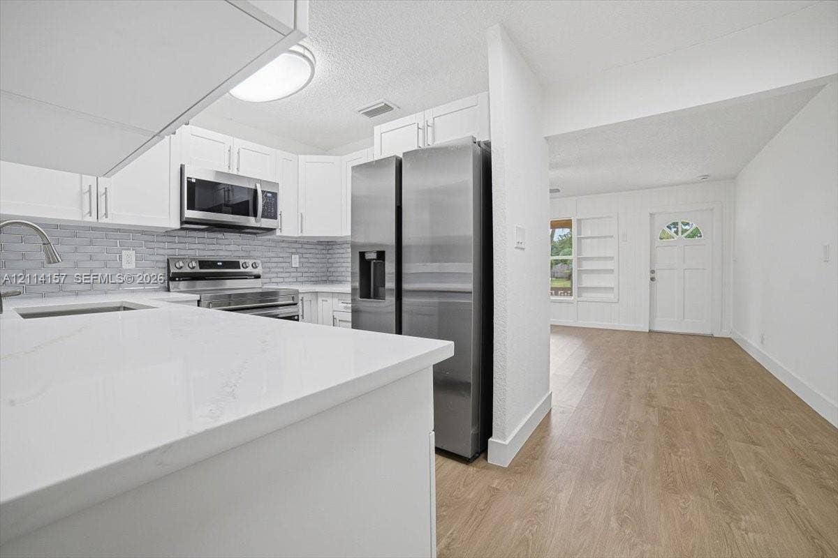 2580 NW 62nd Avenue, Margate, FL 33063 Kitchen featuring white shaker-style cabinetry, solid-surface countertops, a brick-patterned tile backsplash, stainless steel appliances, and wood-finish flooring