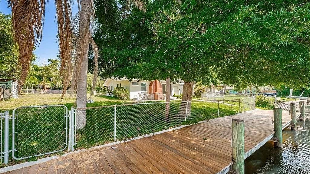 Waterfront property featuring a wood-plank pier, chain-link fencing, expansive lawn, and mature trees