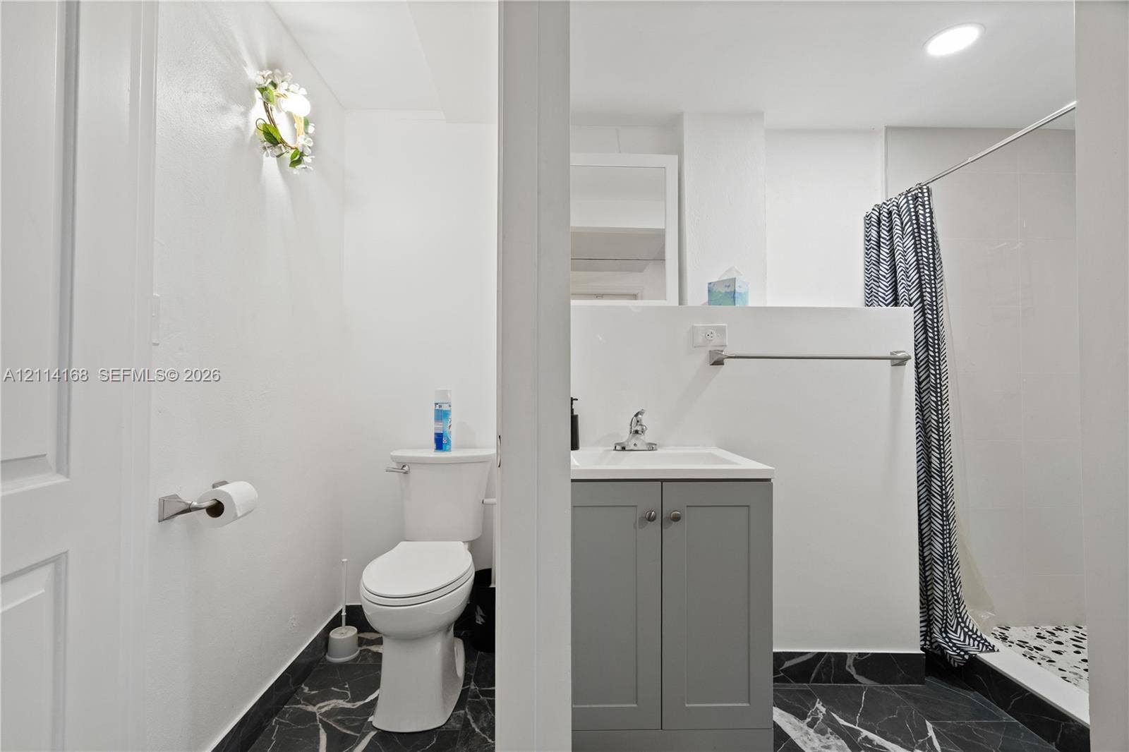 Bathroom featuring a grey vanity with a white countertop, chrome faucet, wall-mounted light fixture, and black veined stone-finish flooring. (Flex space, in-law quarters)