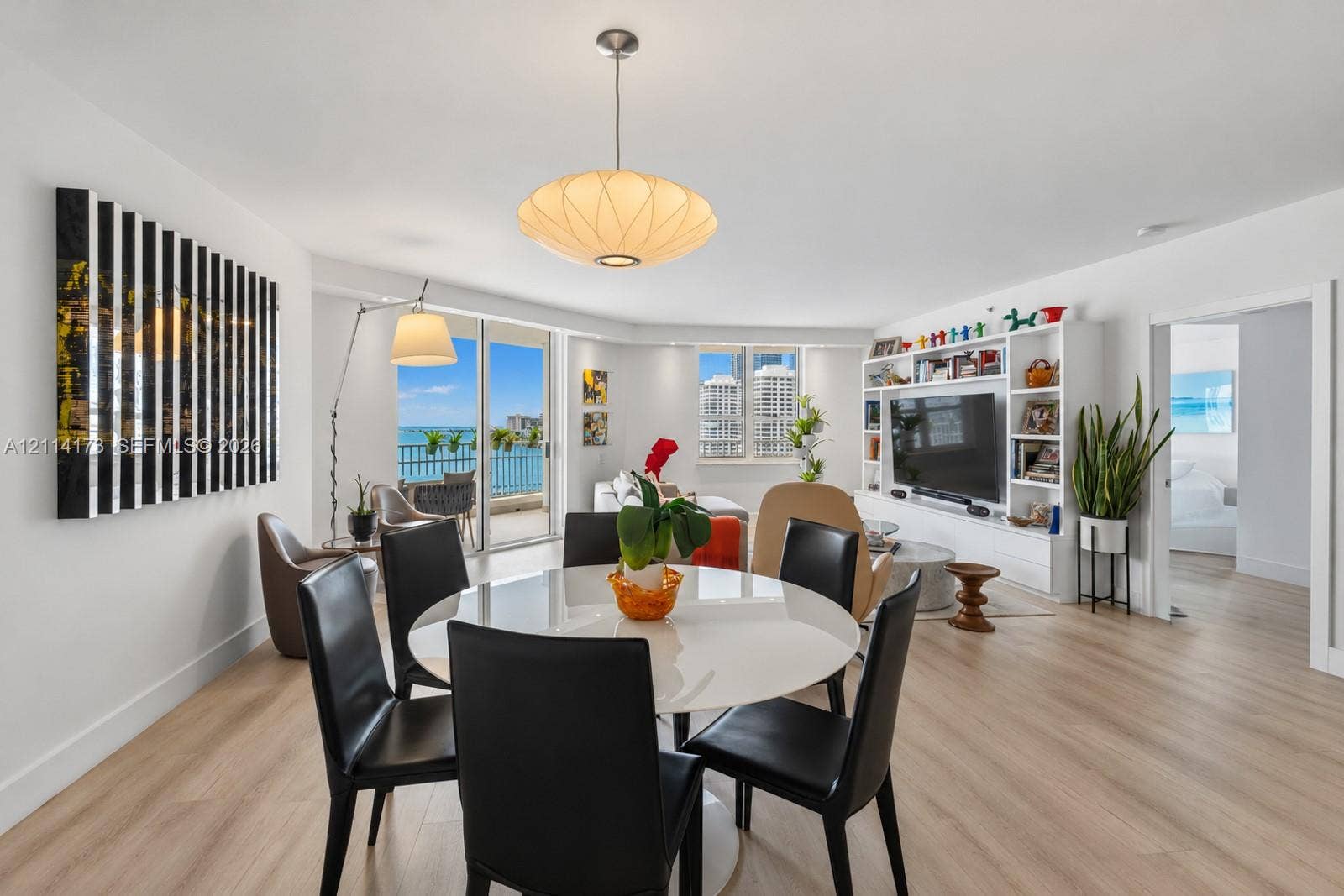 701 Brickell Key Boulevard, #1012, Miami, FL 33131 Open-concept interior featuring light wood-finish flooring, floor-to-ceiling glass sliding doors, a custom built-in media wall with integrated shelving, and a prominent saucer-style pendant light
